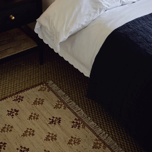 A close-up of a patterned area rug with fringe, styled in a bedroom next to a bed with white and dark navy bedding.