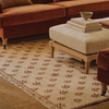 A beige woven area rug with a brown geometric pattern and fringe, styled in a cozy living room setting under an orange sofa and a light wood ottoman.