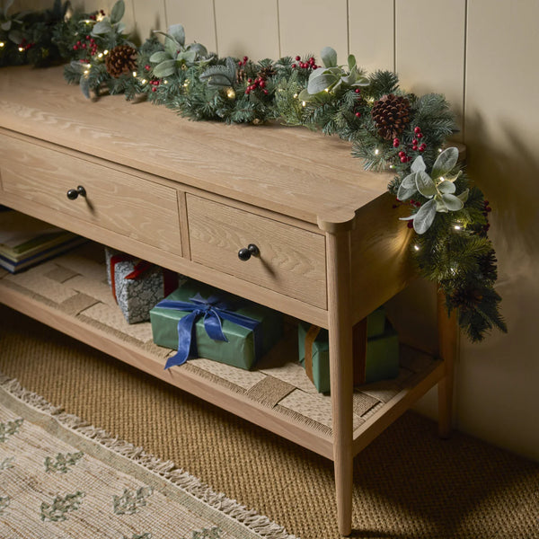 A light natural wood console table styled with a green pine garland, warm string lights, and holiday decorations, with wrapped presents on its lower shelf.