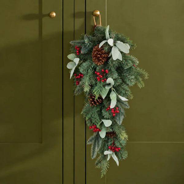 A hanging Christmas garland with green pine branches, red berries, white eucalyptus leaves, and brown pinecones, suspended from a gold hook on an olive green paneled door.