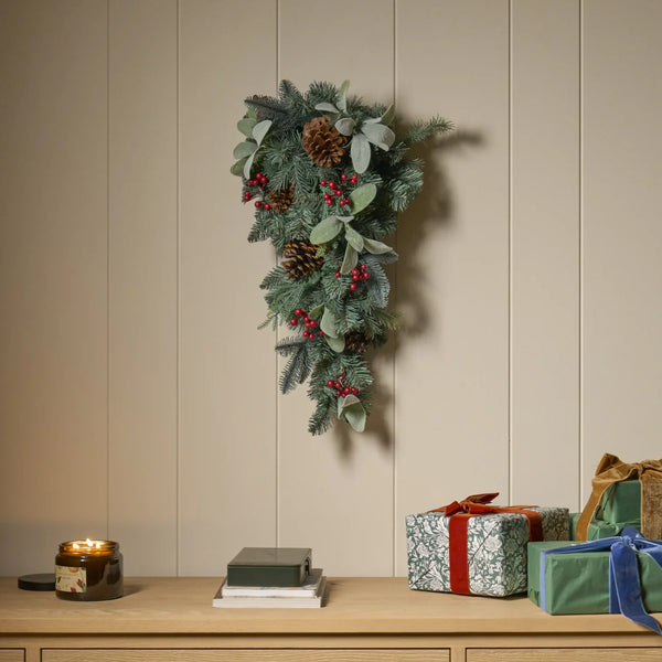 A festive Christmas garland featuring green pine branches, brown pinecones, small red berries, and pale green leaves, hanging on a beige paneled wall above a wooden console table with wrapped gifts and a lit candle.