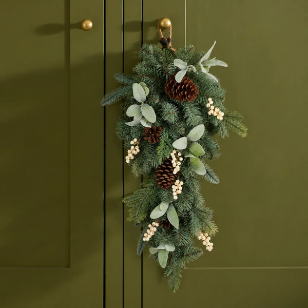 A hanging green garland with pinecones, eucalyptus leaves, and small white berry clusters, displayed against an olive green paneled door.