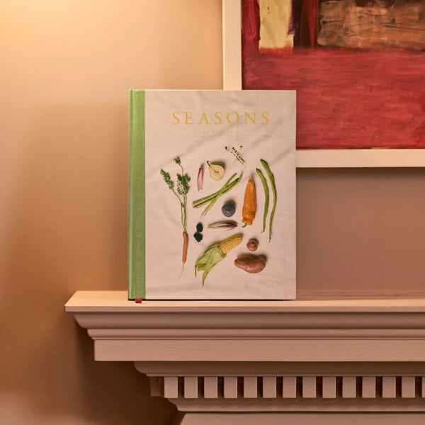 A hardcover book titled 'SEASONS' with a light cover and green spine, standing on a white mantelpiece against a warm beige wall. The cover features watercolor illustrations of seasonal vegetables and herbs.