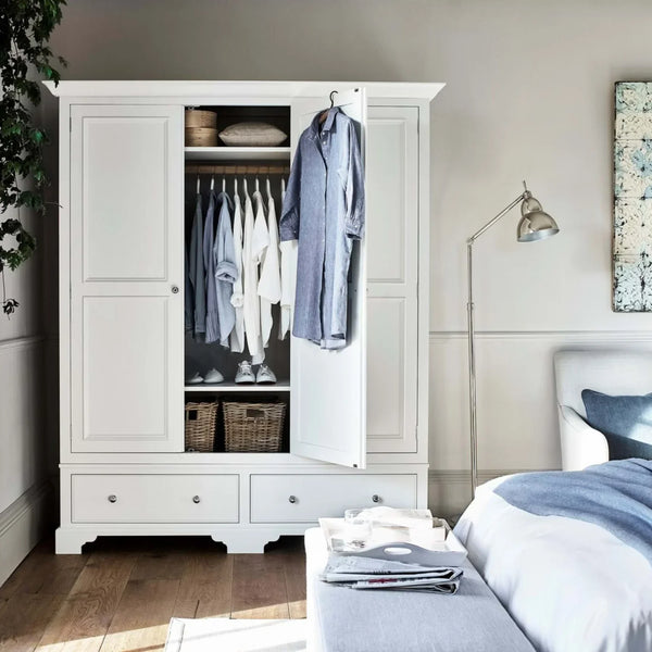 A large white wooden wardrobe with two doors, one open to show hanging clothes and storage baskets, placed in a styled bedroom next to a bed and floor lamp.