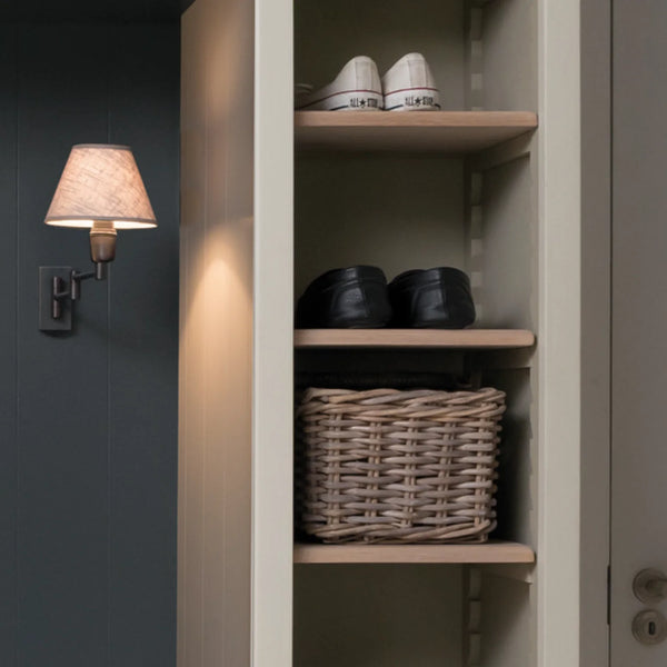 An open interior closet or shelf unit with light wood shelves, containing a pair of white sneakers on the top shelf, black shoes on the middle shelf, and a woven wicker basket on the bottom shelf. A wall-mounted lamp with a beige shade is visible on the left against a dark gray paneled wall.