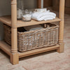 A light natural wood two-tier bathroom shelf with wicker baskets, styled with white towels and a glass jar on a tiled floor.