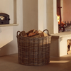 A round woven wicker basket filled with cut firewood logs, placed on the floor in front of a fireplace in a warm, rustic interior.