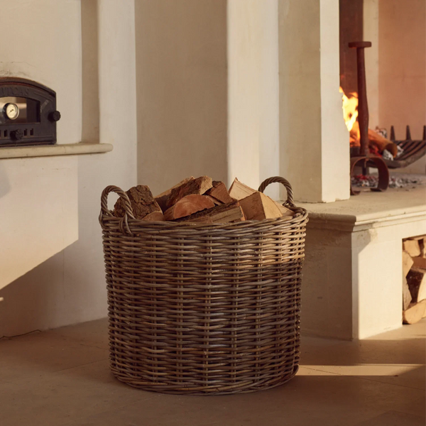 A round woven wicker basket filled with cut firewood logs, placed on the floor in front of a fireplace in a warm, rustic interior.