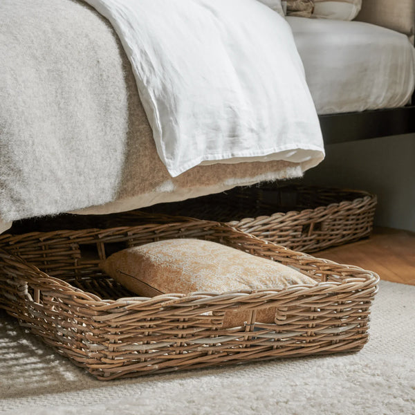 A natural woven wicker storage basket with a slatted base is placed underneath a bed, holding a beige patterned throw pillow. A second basket is visible in the background on a light-colored rug.