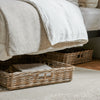 Natural woven wicker storage baskets placed underneath a bed with neutral-toned bedding and a white throw blanket, set on a light rug in a bedroom setting.