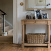 A light natural wood console table with a single drawer and lower shelf, styled with picture frames, a glass jar, and a woven wicker basket in a home entryway.