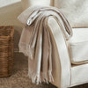 A folded cream-colored throw blanket with fringe trim draped over the arm of a matching upholstered chair, with a woven basket and carpet visible in a softly lit indoor setting.