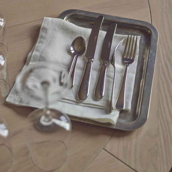 A set of silver-toned flatware, including a spoon, two knives, a butter knife, and a fork, arranged on a folded cream linen napkin atop a rectangular metal tray, placed on a light wood tabletop with blurred wine glasses in the foreground.