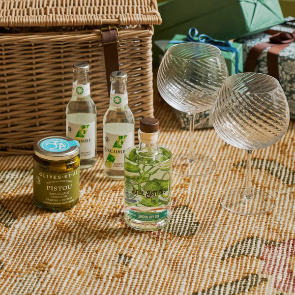 A styled picnic-style product arrangement on a woven blanket with a wicker basket, two clear textured wine glasses, a jar of olive and pistou, and three bottles including a clear bottle labeled 'OLIO sister' and a green-labeled bottle, with wrapped gifts in the background.