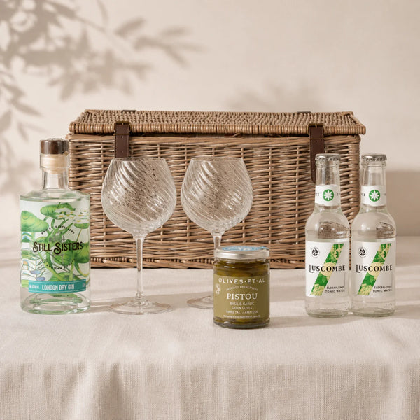 The Gin Hamper with Still Sisters London Dry Gin, two Luscombe Elderflower Tonic Waters, Pistou green olives and two glass goblets, arranged in front of a wicker hamper.