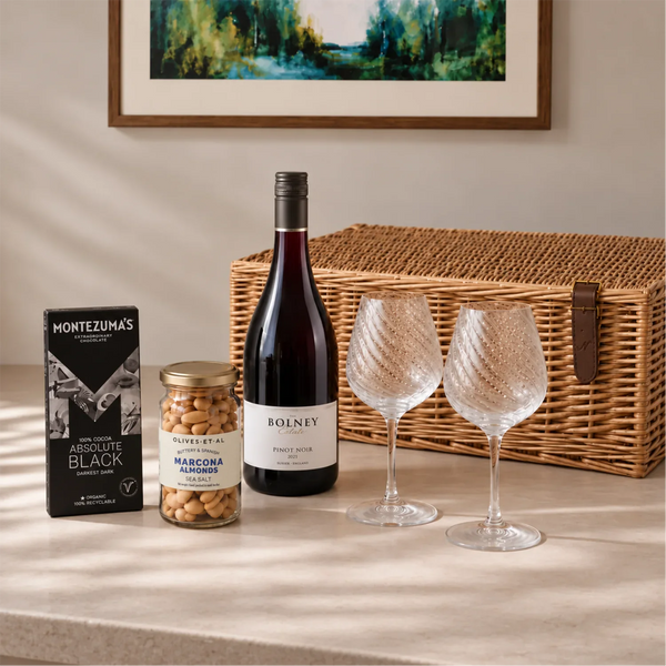 A curated gift set arranged on a tabletop against a warm beige wall, featuring a wicker picnic basket, a bottle of Bolney Pinot Noir red wine, a jar of Macoma Elmsloe nuts, a box of Montezuma's Absolute Black chocolate, and two twisted wine glasses.