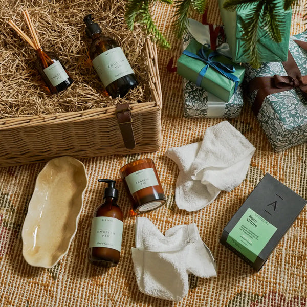 A styled flat-lay of wellness and home products arranged on a woven textile with pine branches and wrapped gifts. Visible items include amber bottles, a candle, white towels, a ceramic dish, and a wicker basket with a reed diffuser bottle.