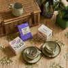A styled flat-lay of Nudi tea products on a woven beige tablecloth, featuring two white tea boxes with purple and pink labels, a purple herbal tea box, and two ceramic teacups and saucers next to a wicker picnic basket.