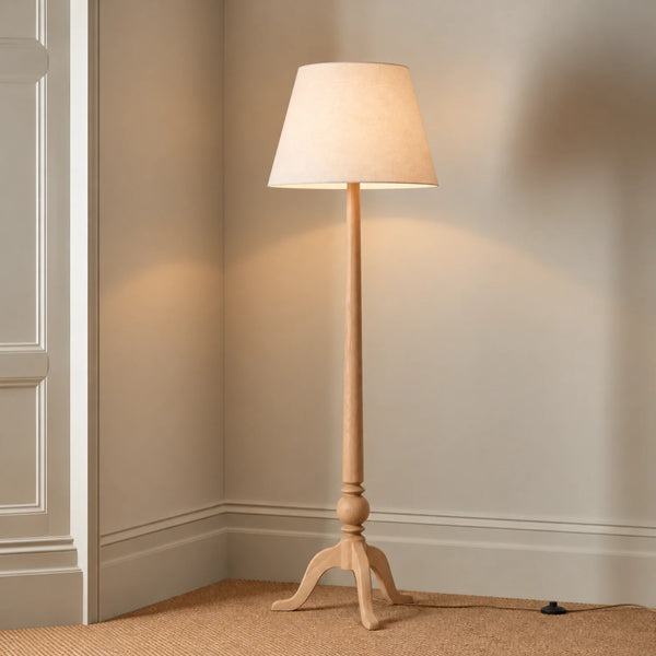 A classic floor lamp with a light natural wood tripod base and a tapered beige fabric drum shade, shown lit in a corner of a room with beige walls and carpet.
