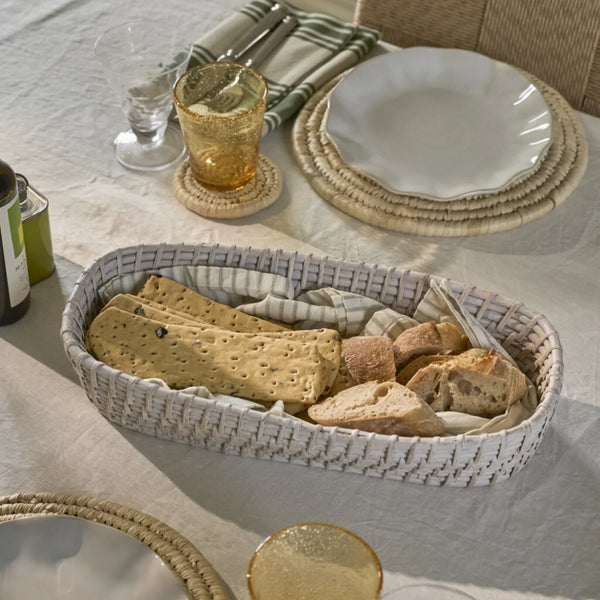 An oval woven bread basket filled with sliced bread and crackers, styled on a white tablecloth at a dining table with plates, glasses, and woven placemats.