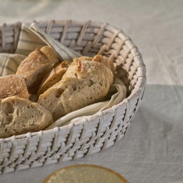 An oval woven bread basket with a white-washed wooden frame, lined with a beige cloth and filled with rustic sliced bread, set on a light fabric surface.