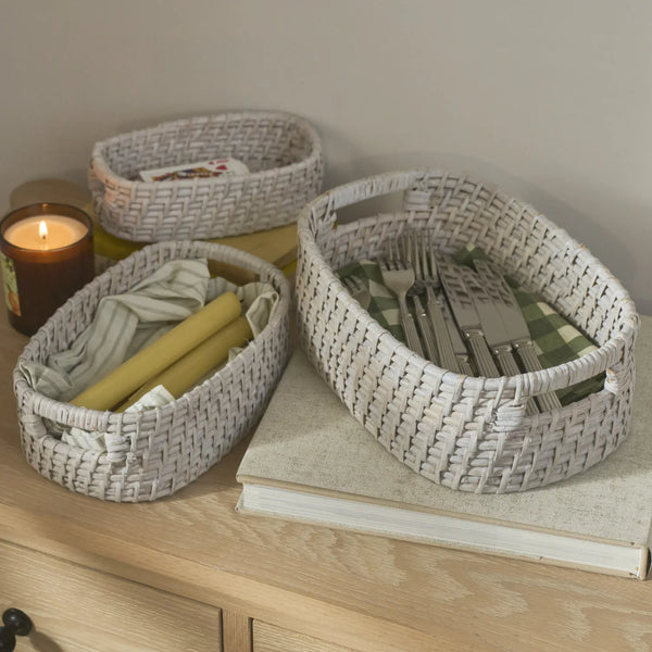 Three oval woven storage baskets in a light natural fiber material, styled on a light wood dresser with a candle, book, and tableware inside.