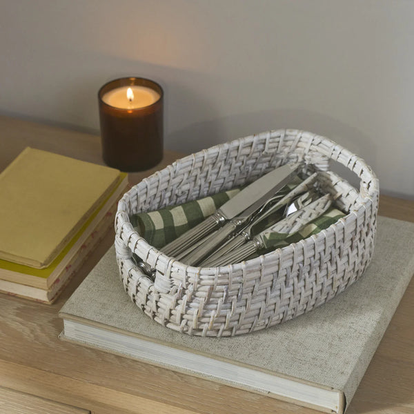 A white woven oval basket holding silver cutlery and green-and-white striped napkins, styled on top of a book on a wooden table with a lit candle and books in the background.