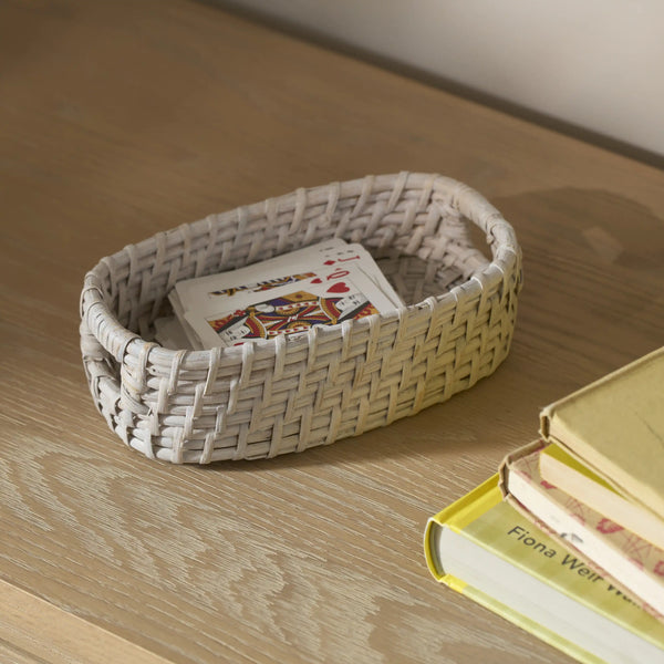 A small woven oval basket in a light natural beige color sits on a wooden tabletop, holding a playing card. A stack of books is partially visible on the right side.
