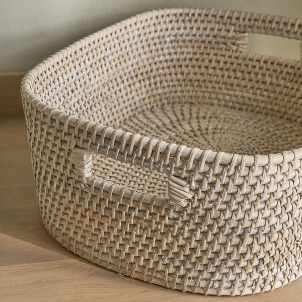 A natural woven rattan storage basket with integrated handles, shown on a light wood floor against a neutral wall.
