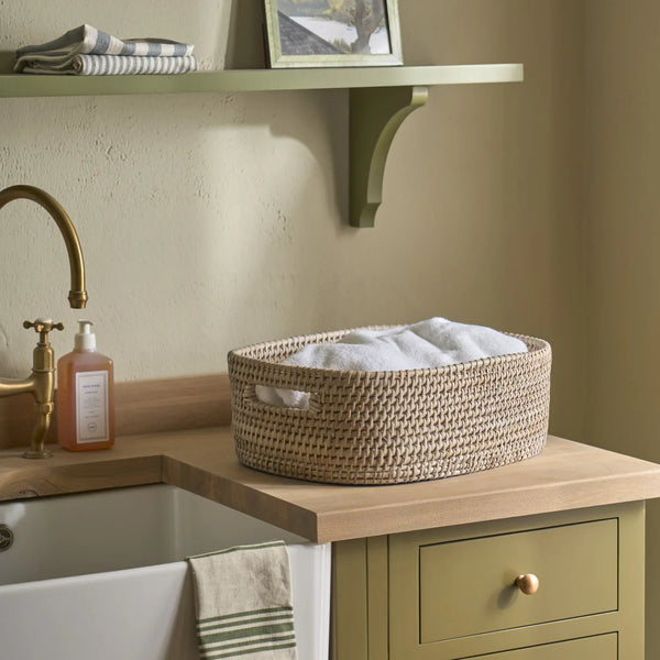 A round woven storage basket with cutout handles sits on a wooden countertop beside a sink in a styled bathroom or utility room. The basket contains folded white towels, and the surrounding environment includes a sage green cabinet, brass faucet, soap dispenser, and shelf.