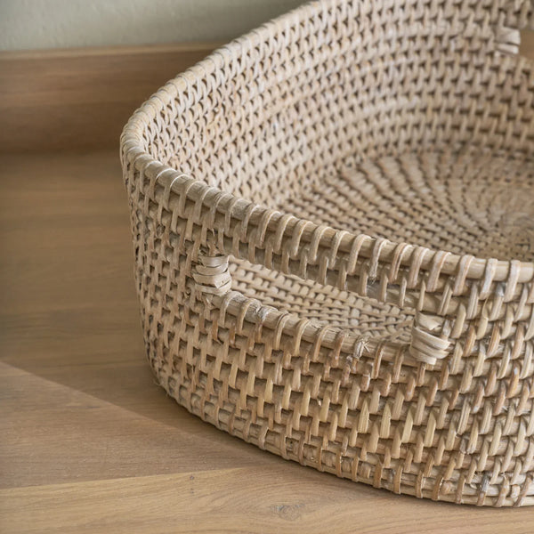A natural woven rattan storage basket with integrated handles, shown on a light wood floor against a neutral wall.