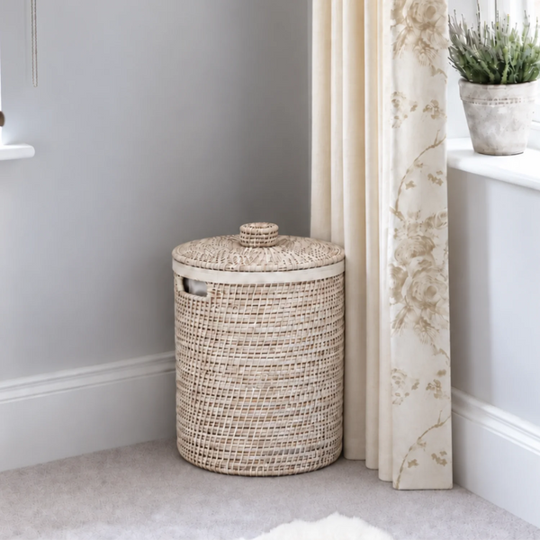 A round woven rattan storage basket with a lid, next to a patterned curtain and windowsill.