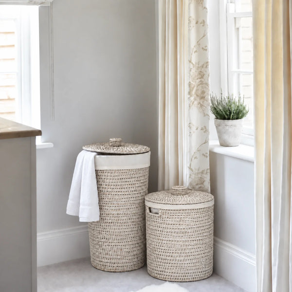 Two woven natural-fiber laundry hamper baskets with lids, one tall and one short, placed in a bright room beside a window with patterned cream curtains and a small potted plant.