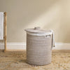 A woven cylindrical laundry hamper with a matching lid, shown in a styled room setting with a beige wall, patterned rug, and a wooden towel rack on the left.
