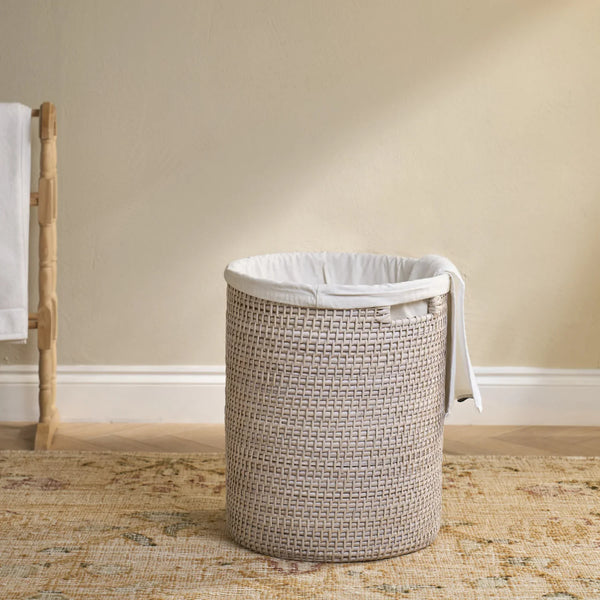 A woven cylindrical laundry hamper basket with a white fabric liner, set on a patterned rug against a beige wall.
