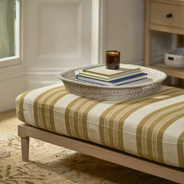 A woven rounded square tray holding stacked books and a small amber candle sits on a light wood bench with cream and tan striped upholstery in a softly lit room.