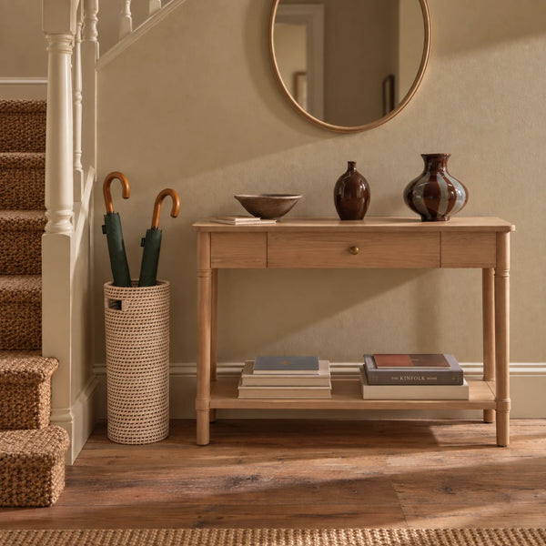 A woven cylindrical umbrella stand holding two closed umbrellas with dark green bodies and warm brown handles, placed beside a light wood console table in a styled interior with carpeted stairs and a beige wall.