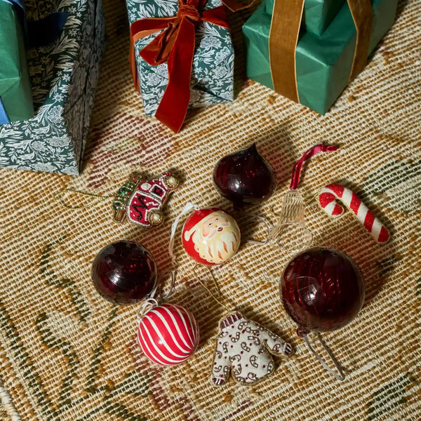A collection of Christmas ornaments arranged on a patterned woven rug, with wrapped gift boxes in the background. The ornaments include dark red glittered balls, a Santa face ornament, a red-and-white striped ball, a small patterned boot ornament, a candy cane, and a small holiday figure.