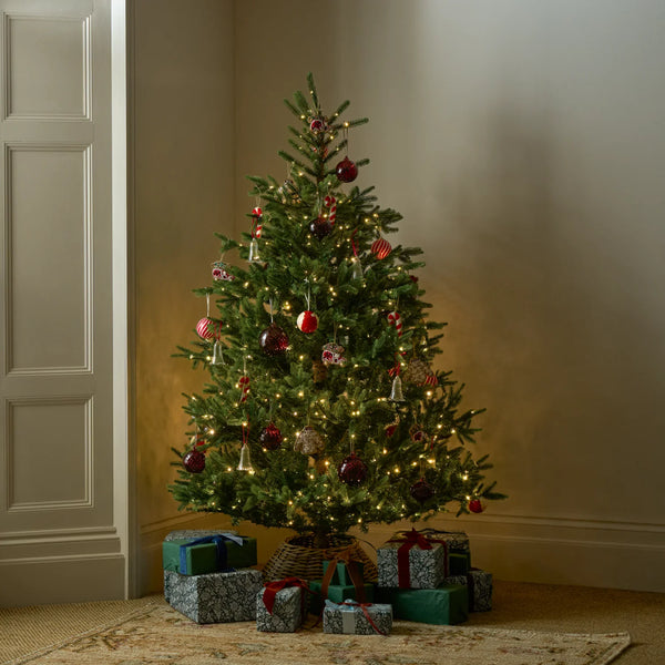 A decorated Christmas tree stands in a warmly lit room corner with wrapped gifts underneath. The tree is green with warm white string lights and red, gold, and silver ornaments.