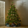 A decorated Christmas tree stands in a corner of a room with warm-toned walls, a door on the left, and wrapped gift boxes at its base. The tree is green with warm white string lights and ornaments in red, pink, silver, and dark tones.