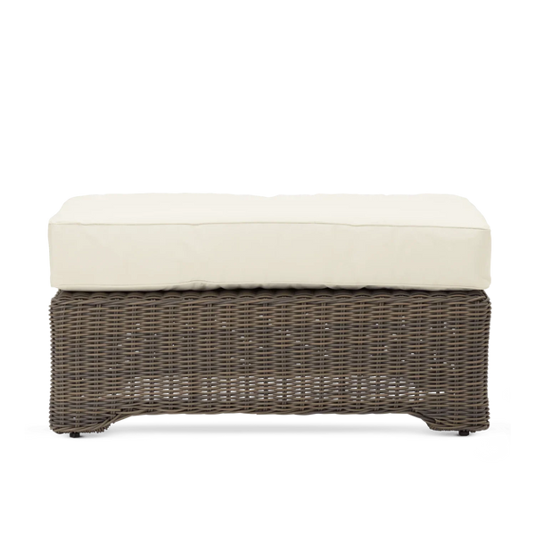 A rectangular outdoor wicker bench or ottoman with a dark brown woven base and thick cream-colored cushions on top, shown against a plain white background.