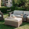 Outdoor wicker patio furniture set on a grassy lawn, featuring a chaise lounge with light beige cushions, a matching ottoman with a decorative tray, and a glass vase on a wooden garden bench in the background.