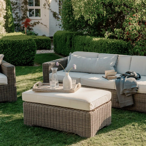 Outdoor wicker patio furniture set on a grassy lawn, featuring a chaise lounge with light beige cushions, a matching ottoman with a decorative tray, and a glass vase on a wooden garden bench in the background.