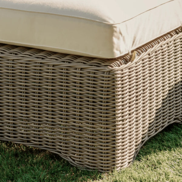 Close-up of a woven wicker outdoor ottoman or bench with a thick cream cushion, shown on green grass outdoors.