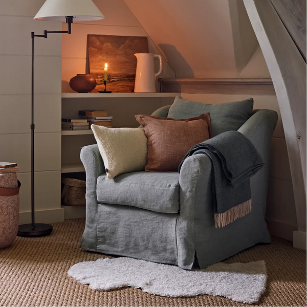 A sheepskin rug, dusty blue-gray upholstered armchair with matching throw pillows and a draped throw blanket, styled in a cozy attic reading nook with a floor lamp, bookshelves, and a slanted ceiling.
