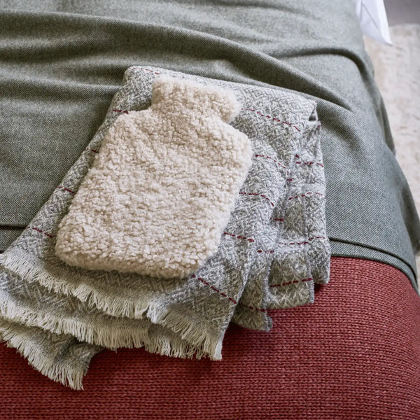A folded cream sherpa hot water bottle cover resting on a textured gray throw blanket with fringe, which is laid over a rust-red upholstered surface.