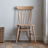 A light natural wood spindle-back chair positioned against a textured off-white wall on a rustic tiled floor, with a woven basket partially visible on the left.