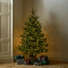 A decorated artificial Christmas tree with warm white string lights stands in a corner of a room, surrounded by wrapped gift boxes on the floor.