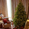 A lit Christmas tree stands in a decorated living room beside a beige armchair with a red pillow, wrapped gifts under the tree, and tall patterned curtains in the background.
