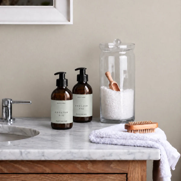 Two amber pump bottles of English Fig Soap hand and body wash with black caps, styled on a white marble bathroom counter next to a glass jar of bath salts and a wooden brush on a white towel.